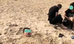 男子把自己埋进沙里，把头露在外面，宠物狗在他头上跳来跳去